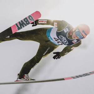 A professional ski jumper soars gracefully through the air during a competition, captured against a bright winter sky.