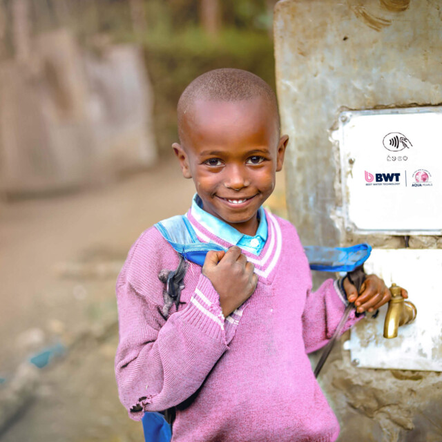 A smiling child stands in front of a tap at a water station, supported by BWT.